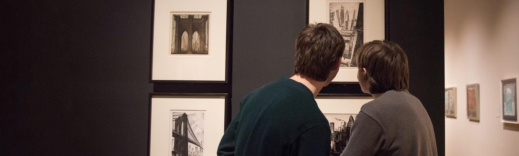 The backs of two visitors, who are looking closely at black and white artworks mounted on a dark wall in an exhibition gallery