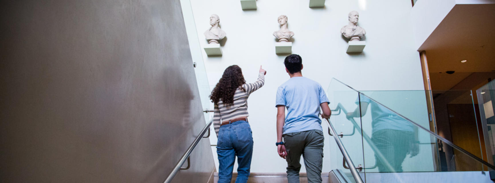 View from behind to museum visitors ascending a staircase, with busts on the wall in front of them