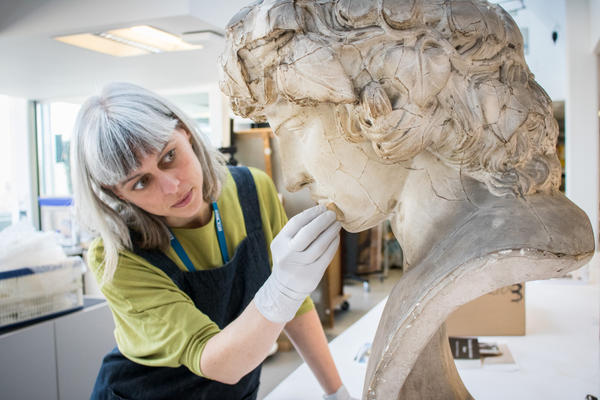 A woman in a green top and dark overalls examines a large bust of the head of Greek boy Antinous