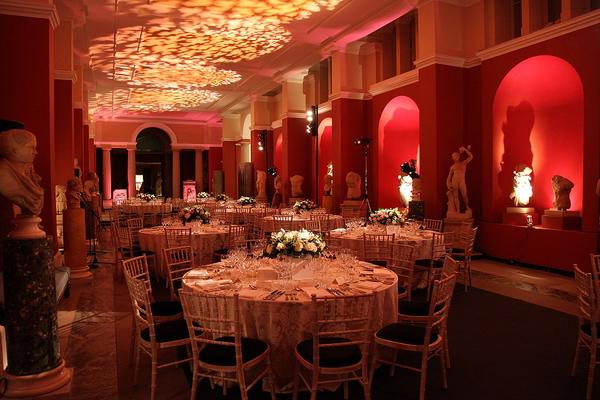 Long museum gallery, lined with sculptures and lit in red and white, set up for a formal dinner