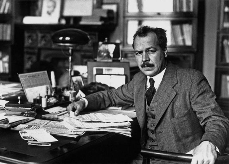 Black and white picture of a men sitting at his desk in a library or study, letters atop a newspaper