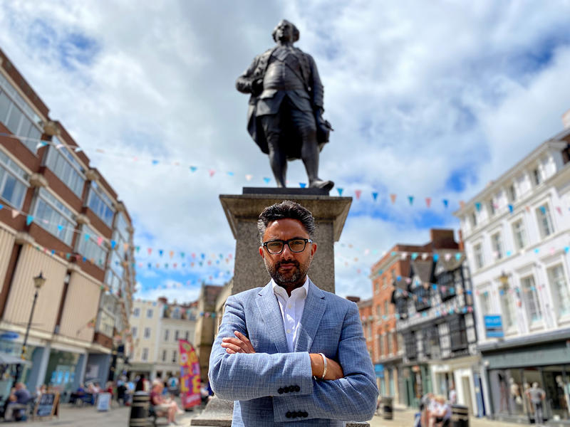 Colour photo of Sathnam Sanghera standing in front of a monument flanked by buildings, with flag bunting over the streets