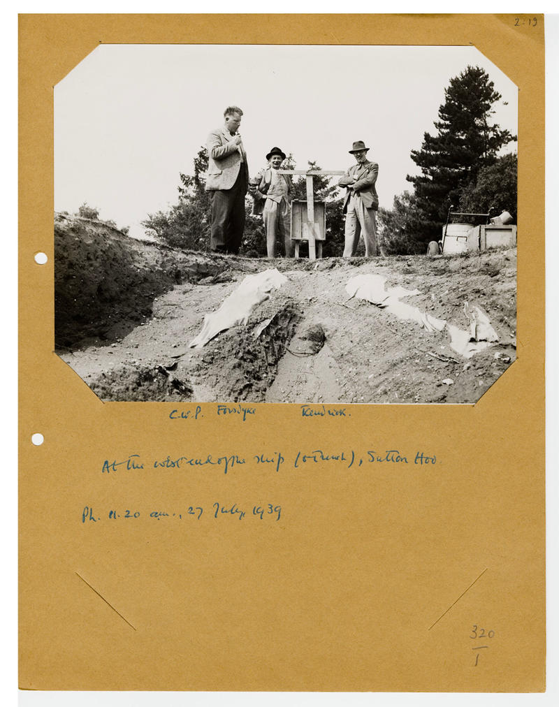 A black and white photograph of figures next to an archaeological dig, with handwritten notes underneath
