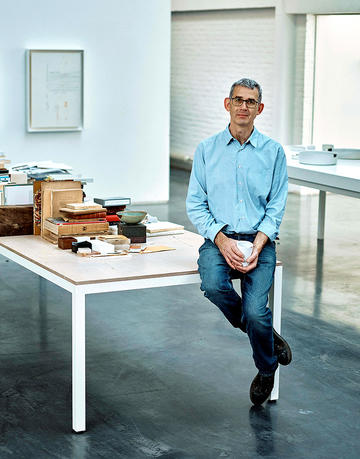 Edmund de Waal sitting on a white table piled with books holding a cup of coffee wearing blue shirt and jeans