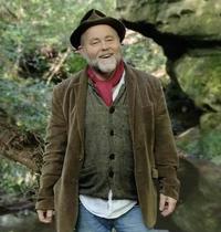 Full length portrait picture of David Tong, wearing a green hat and long coat, gilet, jeans and a red scarf, he is standing in nature with rocks and greenery behind him